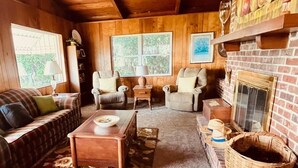 Interior acogedor de una cabaña con techo de madera, chimenea, sofá, una mesita de centro y ventana con vistas a follaje.
