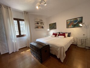 A bedroom with a bed, a chest of drawers, a bedside table, a lamp, a window with curtains, and a bookshelf.