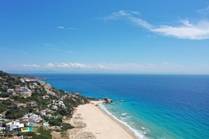 Vista costera con playa arenosa, mar azul cristalino y una colina con edificios.