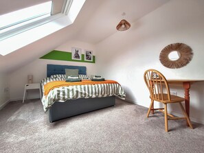 Ein Schlafzimmer mit schrägverdeckter Decke, einem Bett mit gestreifter Bettwäsche, einem Schreibtisch mit Stuhl und einem wandmontierten Spiegel.