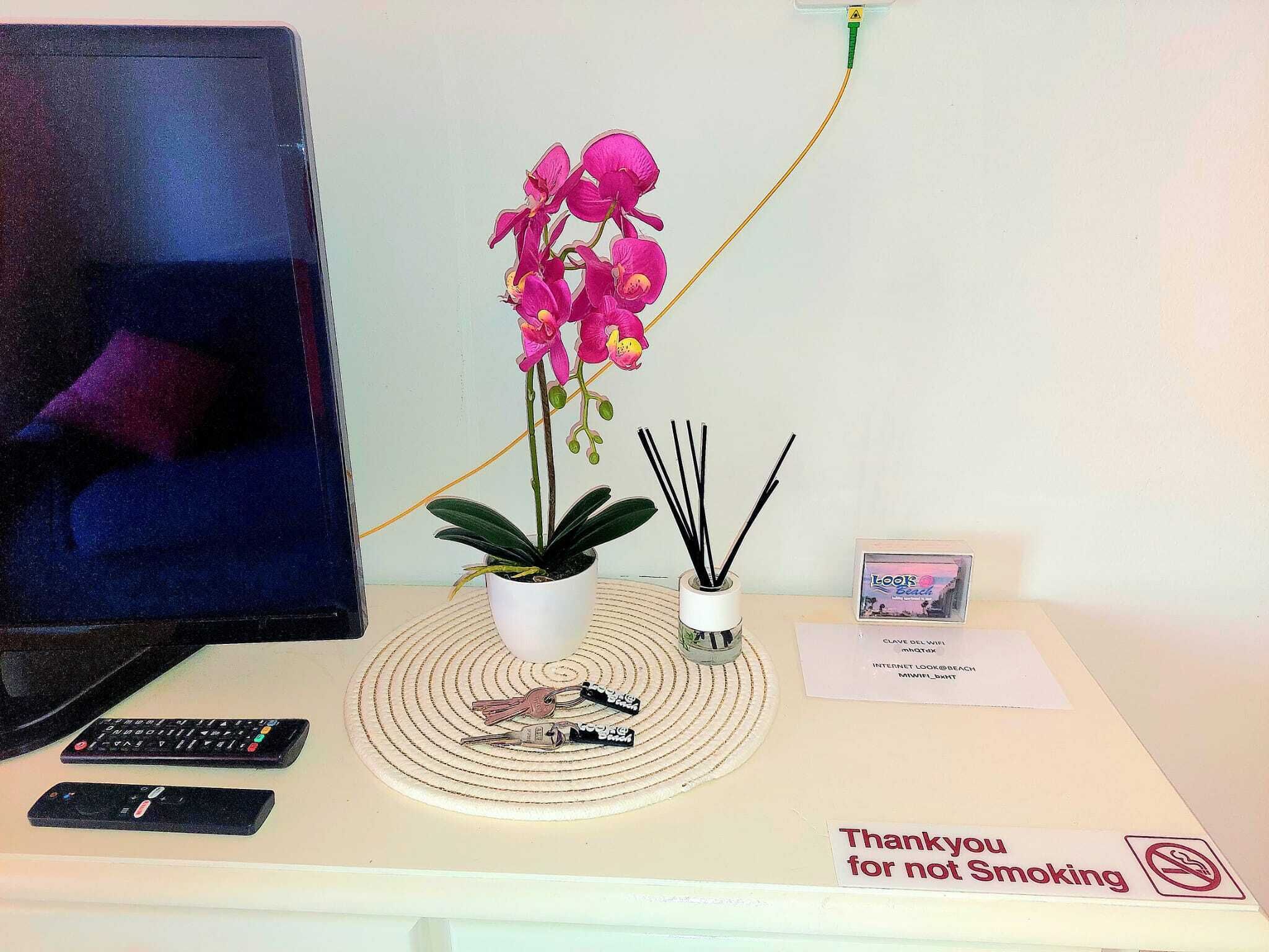 Ein Schreibtisch mit einem Computermonitor, zwei Fernbedienungen, einer Topfpflanze mit Orchideen, einem Diffusor mit Rattanstäbchen, einer gerahmten Karte und einem Schild mit der Aufschrift 'Thank you for not Smoking'.