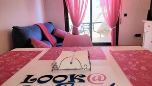 A hotel room with a bed, a desk, a chair, a window with pink curtains, and a view of the outside.