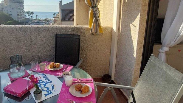 A round glass table with a pink placemat, a chair, a small potted plant, a glass of water, a cup, a plate with food, and a pink notebook on the balcony.