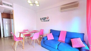 A living room with a blue sofa and pink chairs, a dining table, and a kitchen area.
