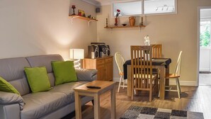 Sala de estar con un sofá gris, cojines verdes, una mesa de centro de madera y un área de comedor con una mesa y sillas de madera. Hay estantes sobre el sofá con objetos decorativos y una ventana con vistas.