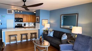 A modern living room with a blue wall, a dark grey sofa, a round coffee table, and a kitchen area with wooden cabinets and a white countertop. - Sugar Beach Oceanfront Condo with Spectacular Views and Steps to Beach (Kihei)