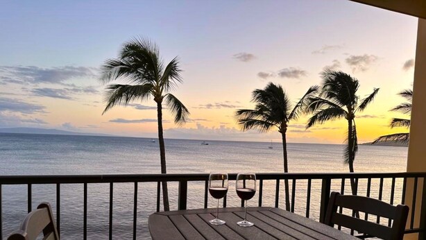 A wooden table with two chairs on a balcony overlooking the sea with palm trees and a sunset. - Sugar Beach Oceanfront Condo with Spectacular Views and Steps to Beach (Kihei)