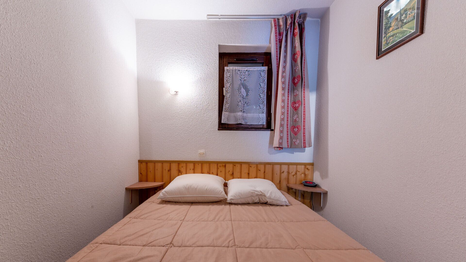 Ein Schlafzimmer mit einem hölzernen Kopfteil, einem Bett mit zwei Kissen, einem Nachttisch, einer Wanduhr und einem Fenster mit gemusterten Vorhängen.