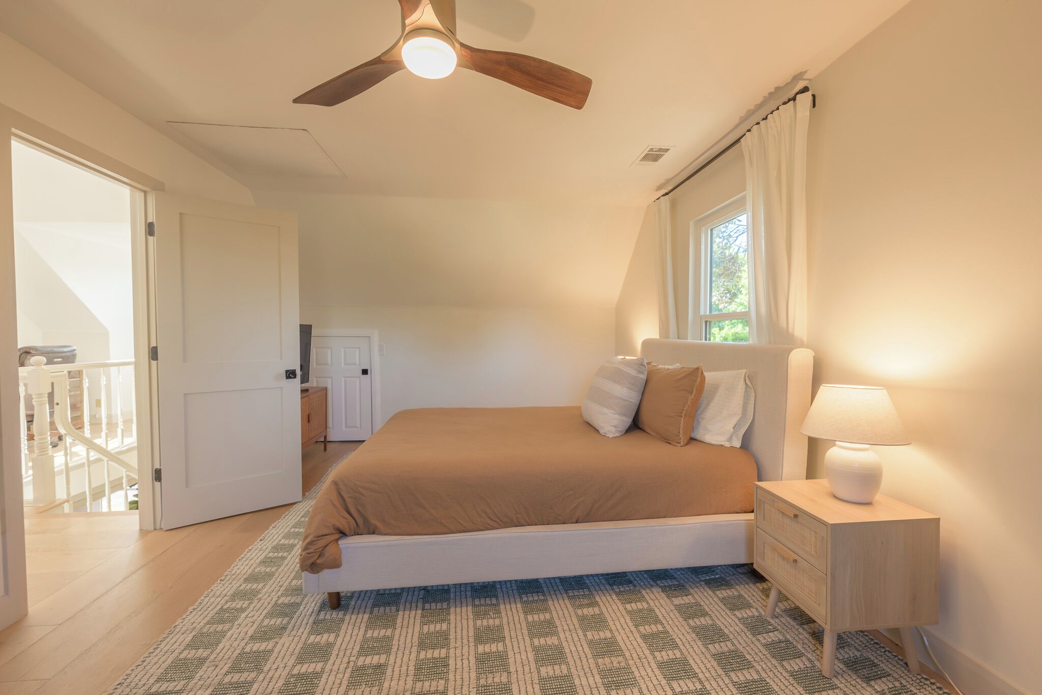 A bedroom with a bed, bedside table, lamp, ceiling fan, and a view of the outdoors.