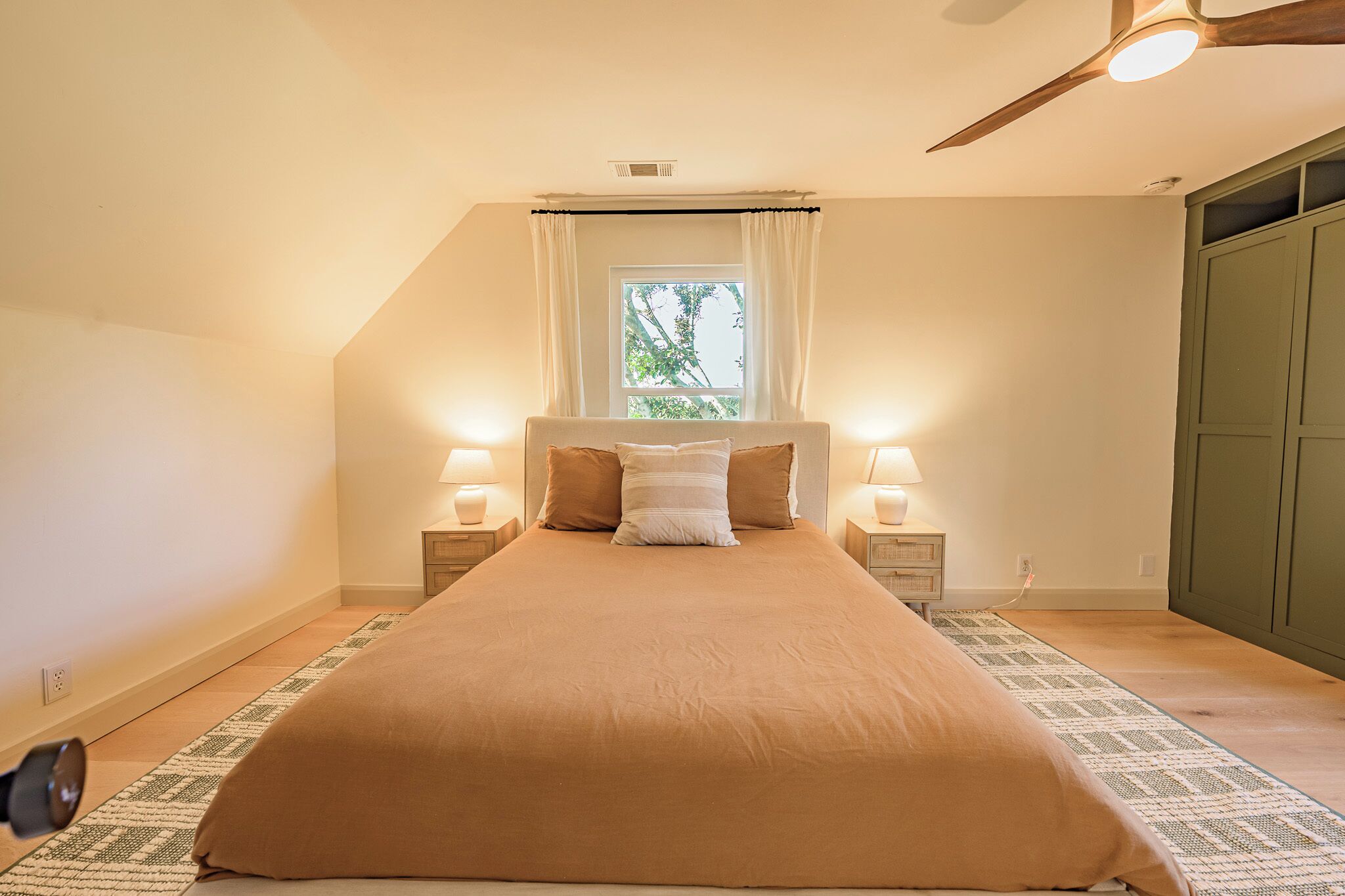 A bedroom with a large bed, two bedside lamps, a window with curtains, and a ceiling fan.