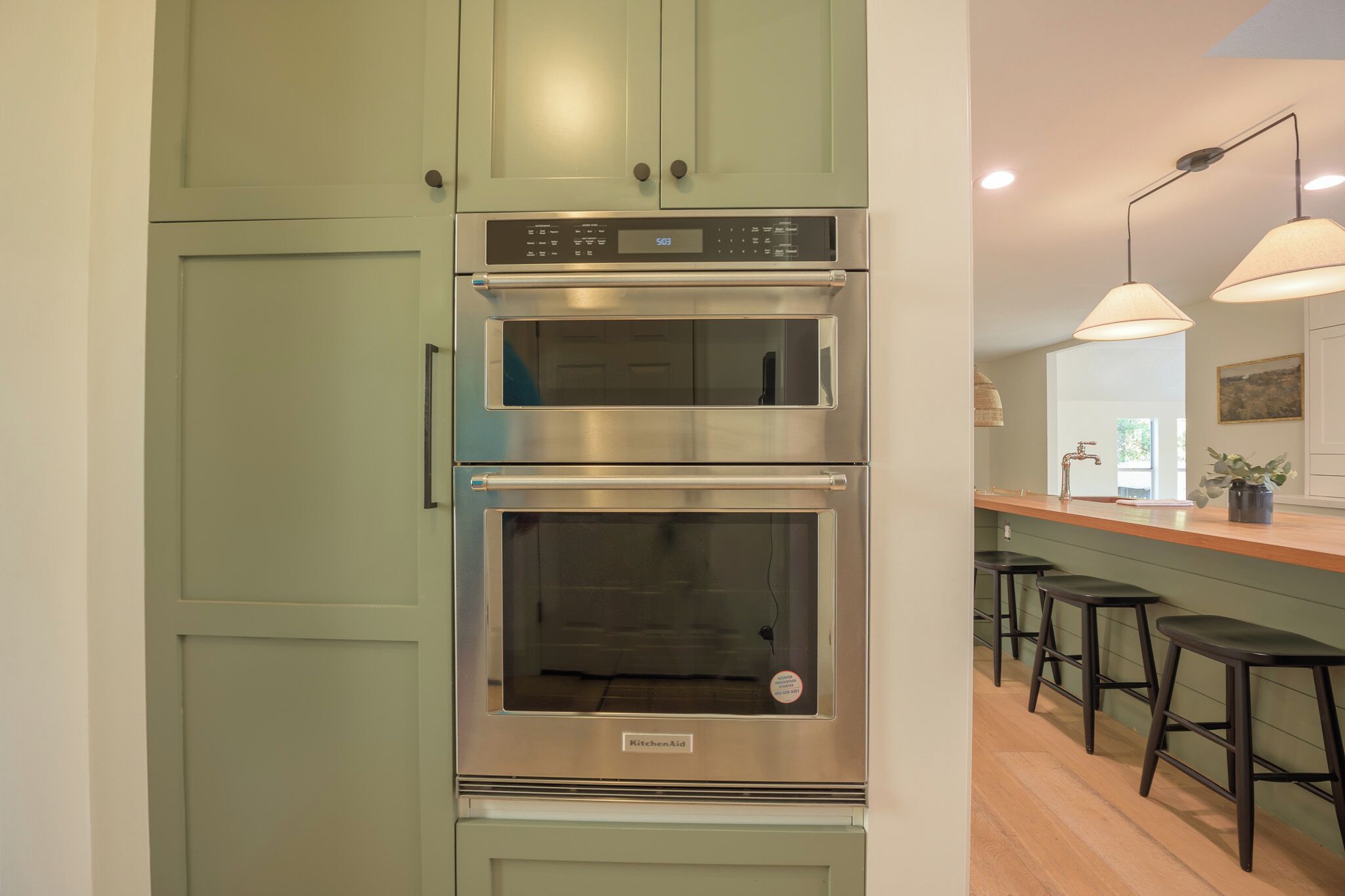 A kitchen with two stainless steel ovens, a green cabinet, a wooden countertop, and black bar stools.