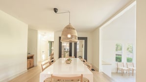 Uma sala de jantar ampla com uma grande mesa branca, cadeiras de vime e uma luminária pendente.
