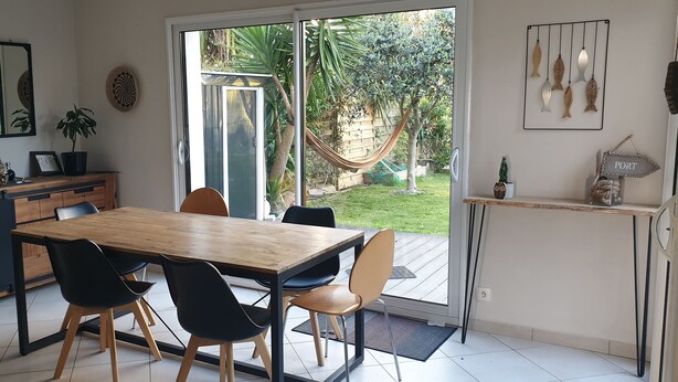 Une cuisine moderne avec une table et des chaises en bois, une grande fenêtre donnant sur un jardin et un petit îlot central doté d’un évier et d’un plan de travail.