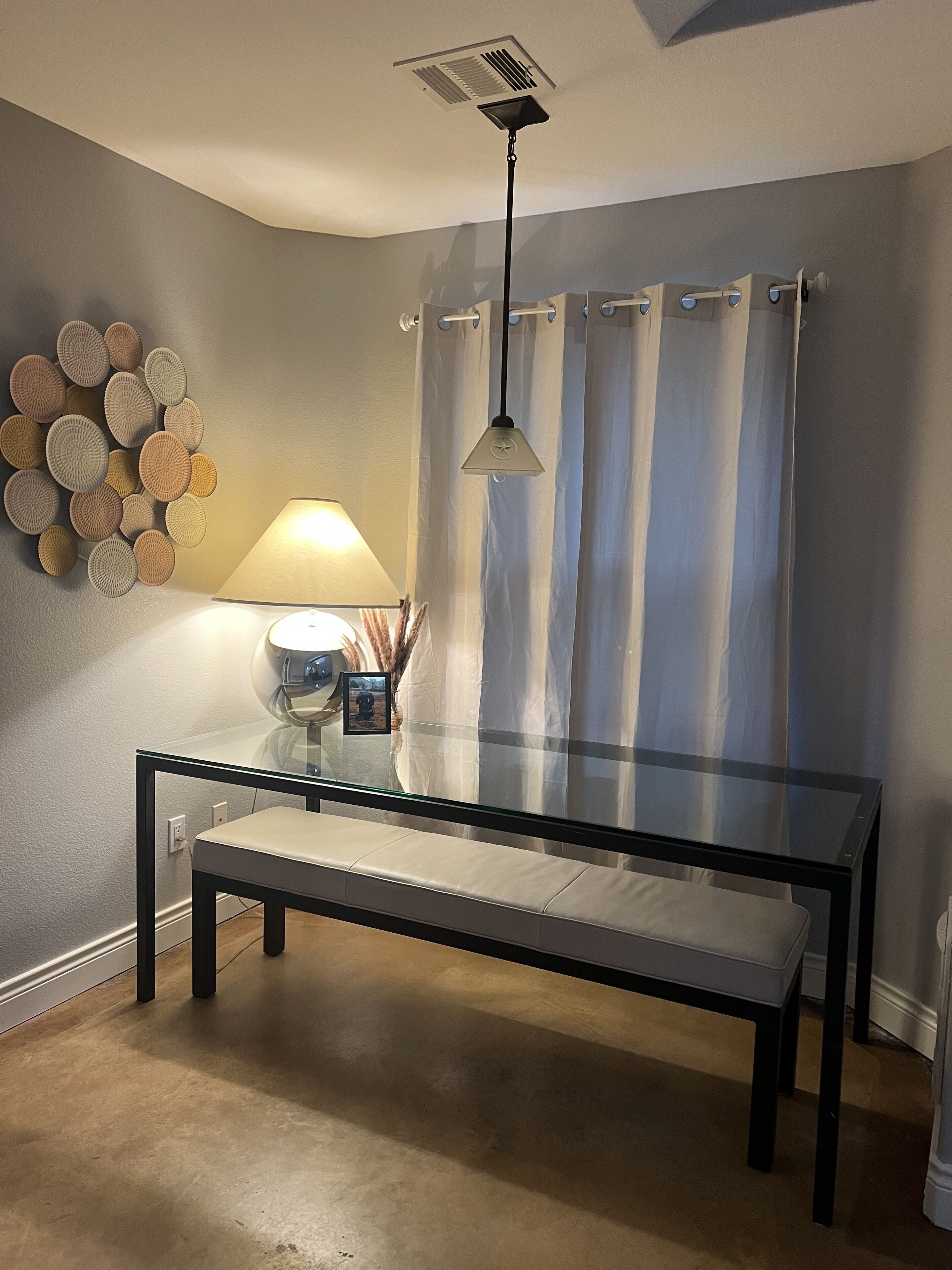 Une pièce avec une table en verre, un banc, une lampe, un luminaire fixé au mur et un rideau.