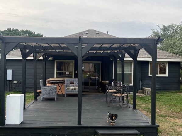Une terrasse extérieure couverte avec une pergola, un coin repas avec des chaises et une table, et un chien debout sur la terrasse.