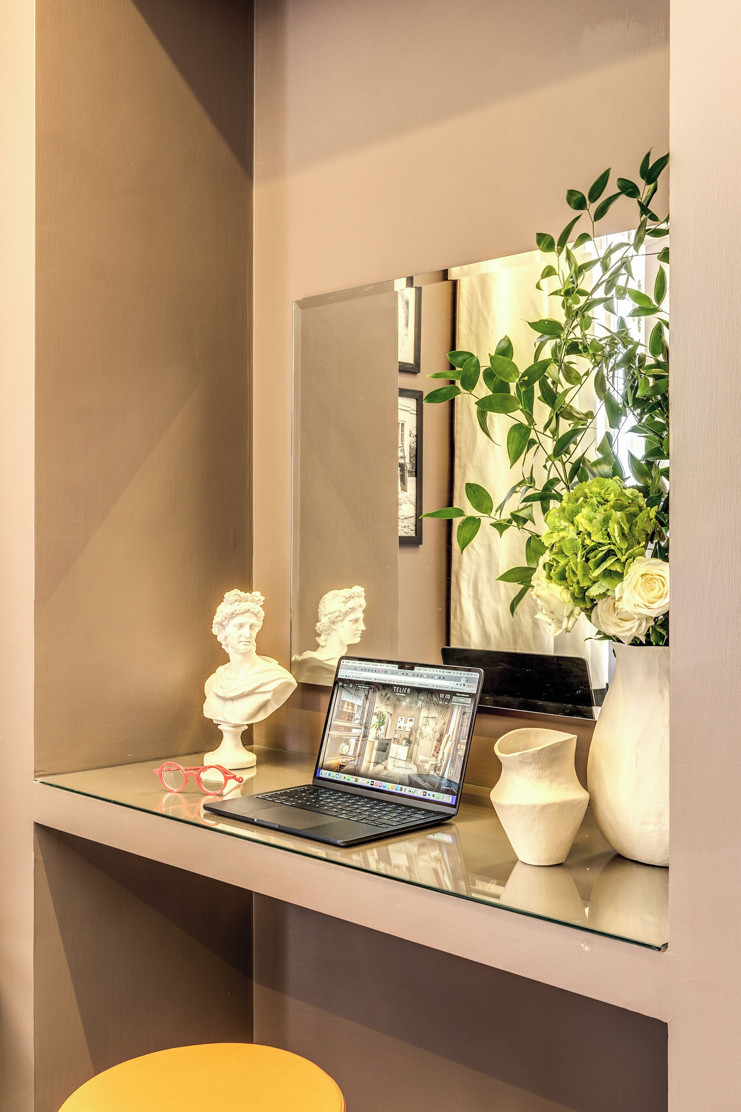 A modern room with a glass desk, a laptop, a bust sculpture, a vase with flowers, and a mirror reflecting the room.