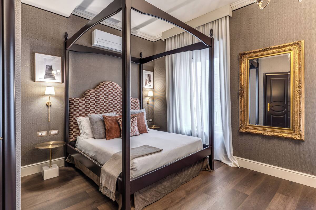 Una cama con dosel, un cabecero con estampado geométrico, lámparas de noche y un espejo grande y ornamentado en la pared.