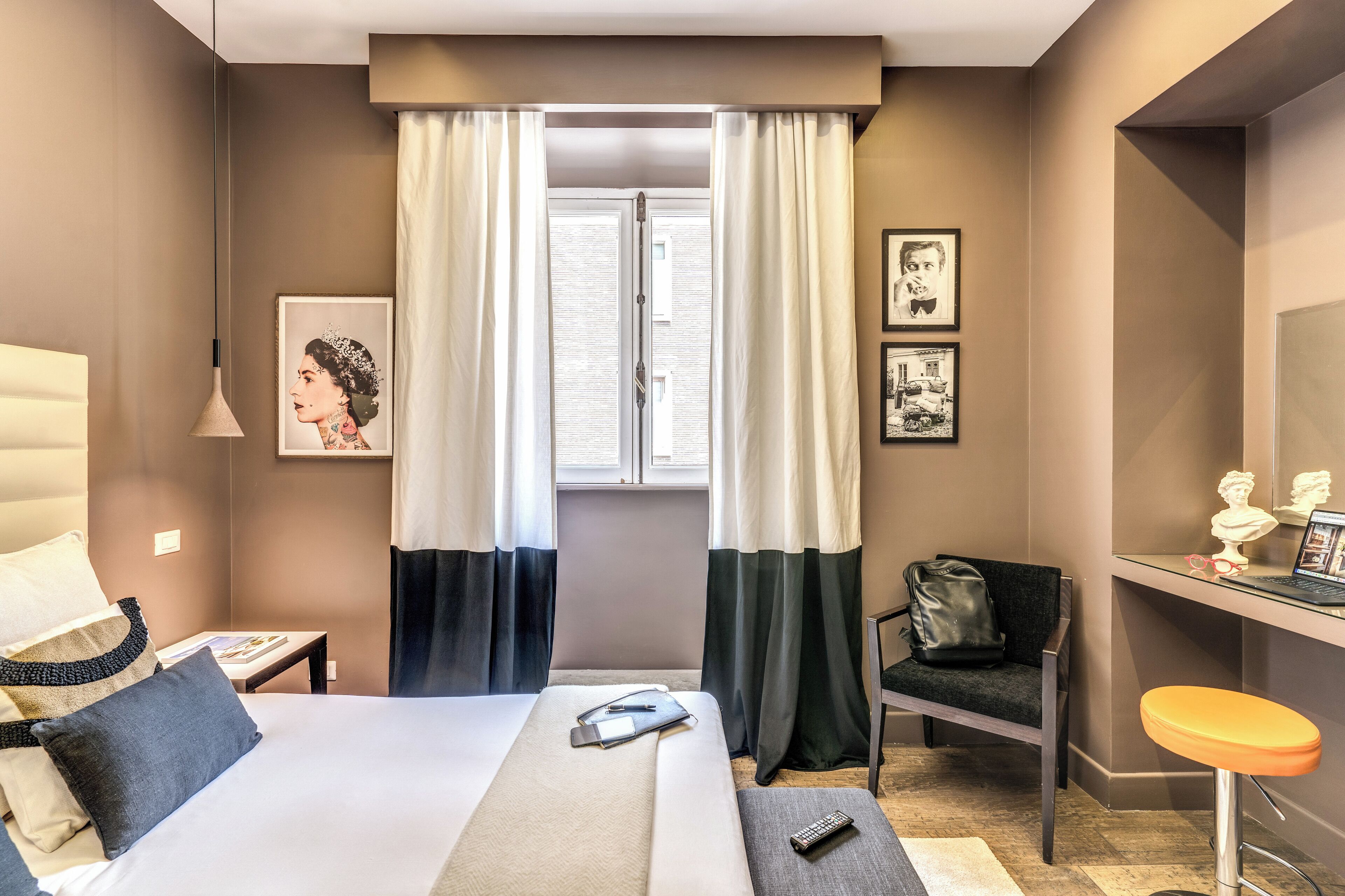 A hotel room with a bed, a desk with a chair, a window with curtains, and artwork on the walls.