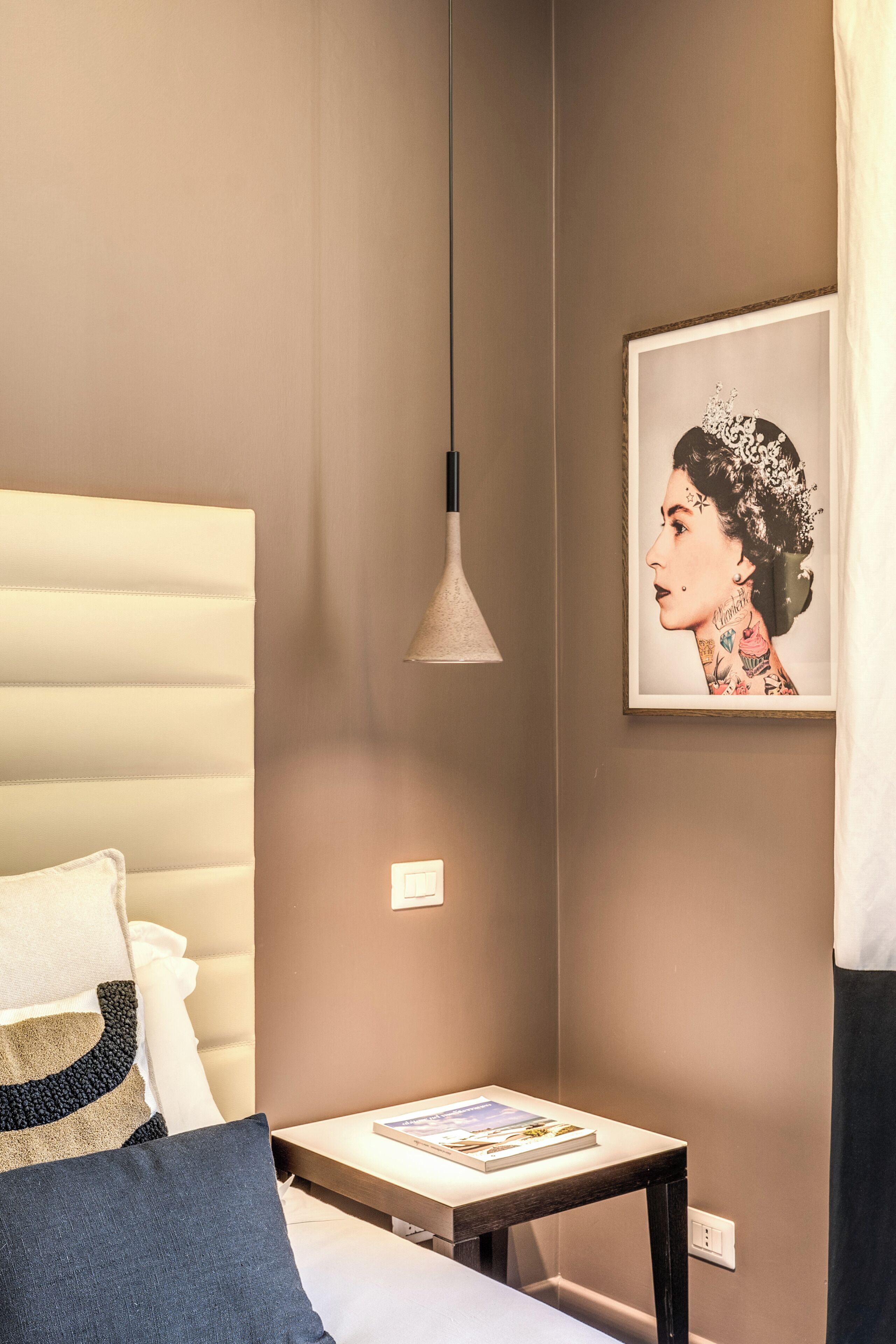 Un dormitorio moderno con cama, mesita de noche con un libro, lámpara de pared y una obra de arte enmarcada.