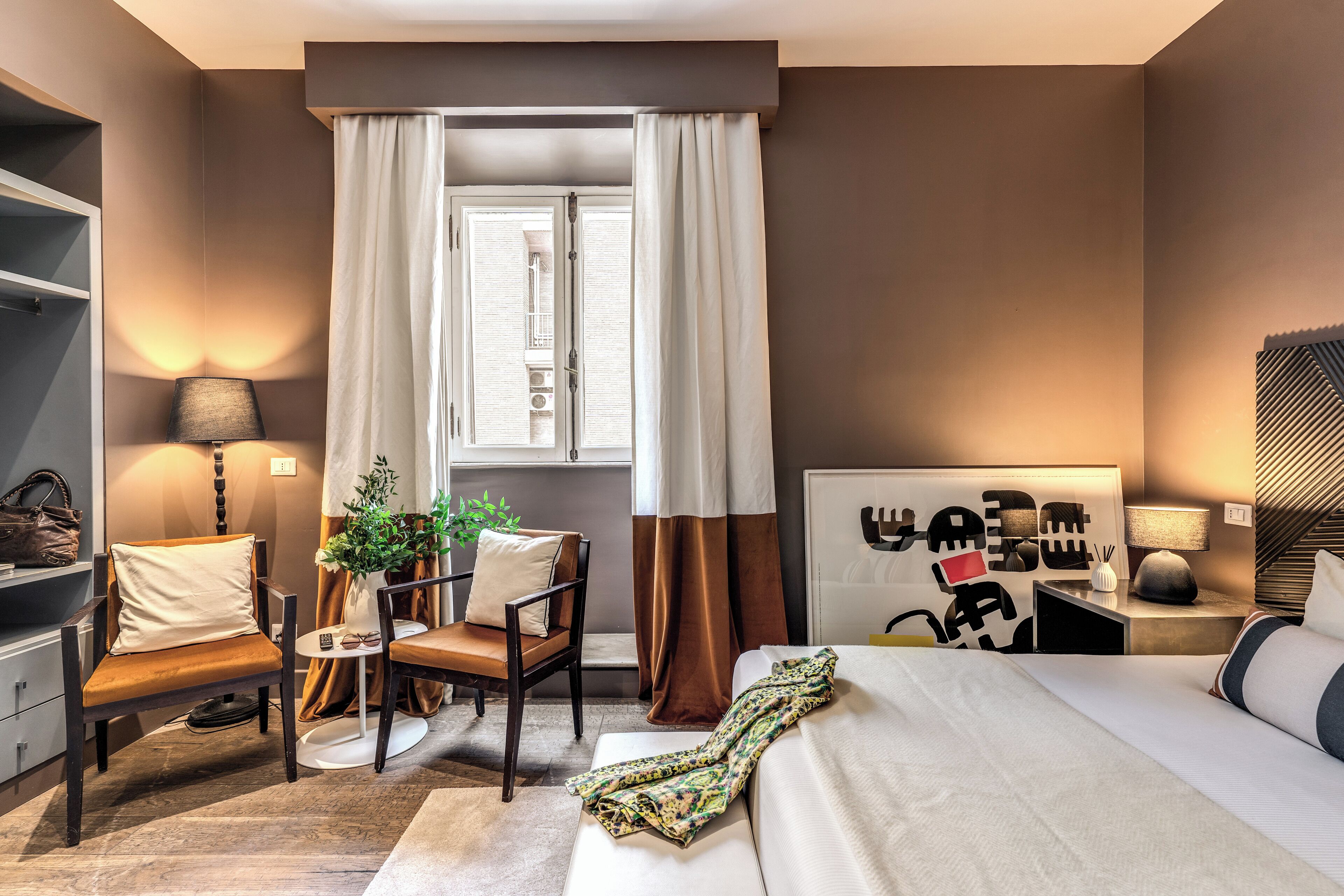 A hotel room with a bed, two chairs, a small table, a lamp, a nightstand, and a window with curtains.