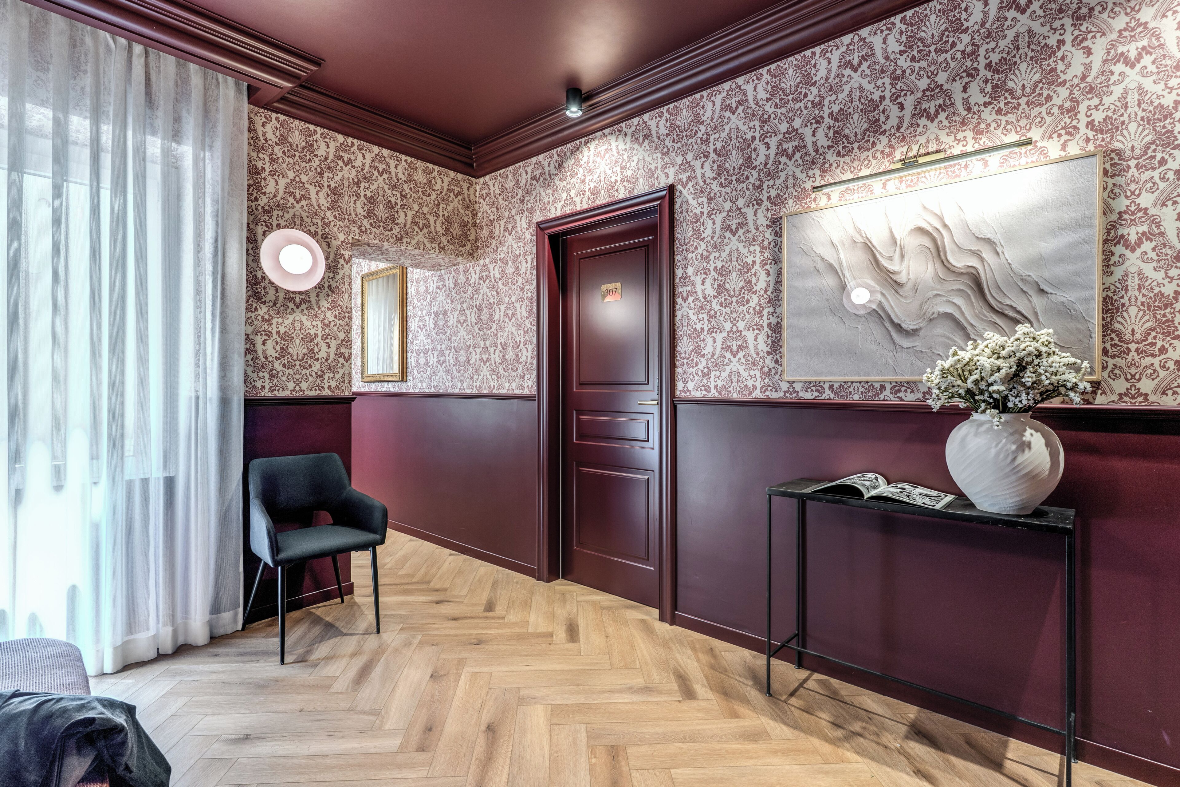 A room with a herringbone floor, a dark wooden door, a black chair, a console table with a vase, and patterned wallpaper.