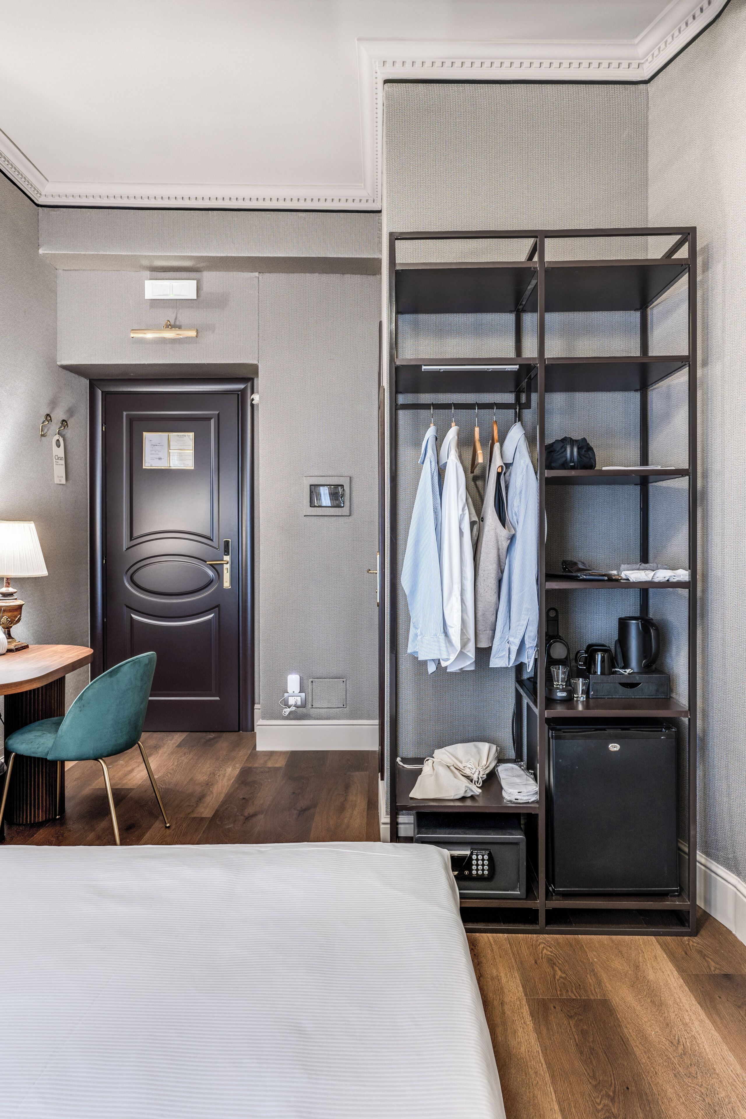 Hotelkamer met een bed, bureau met lamp, stoel, garderobe met kleding, kleine koelkast en nachtkastje met telefoon en beker.