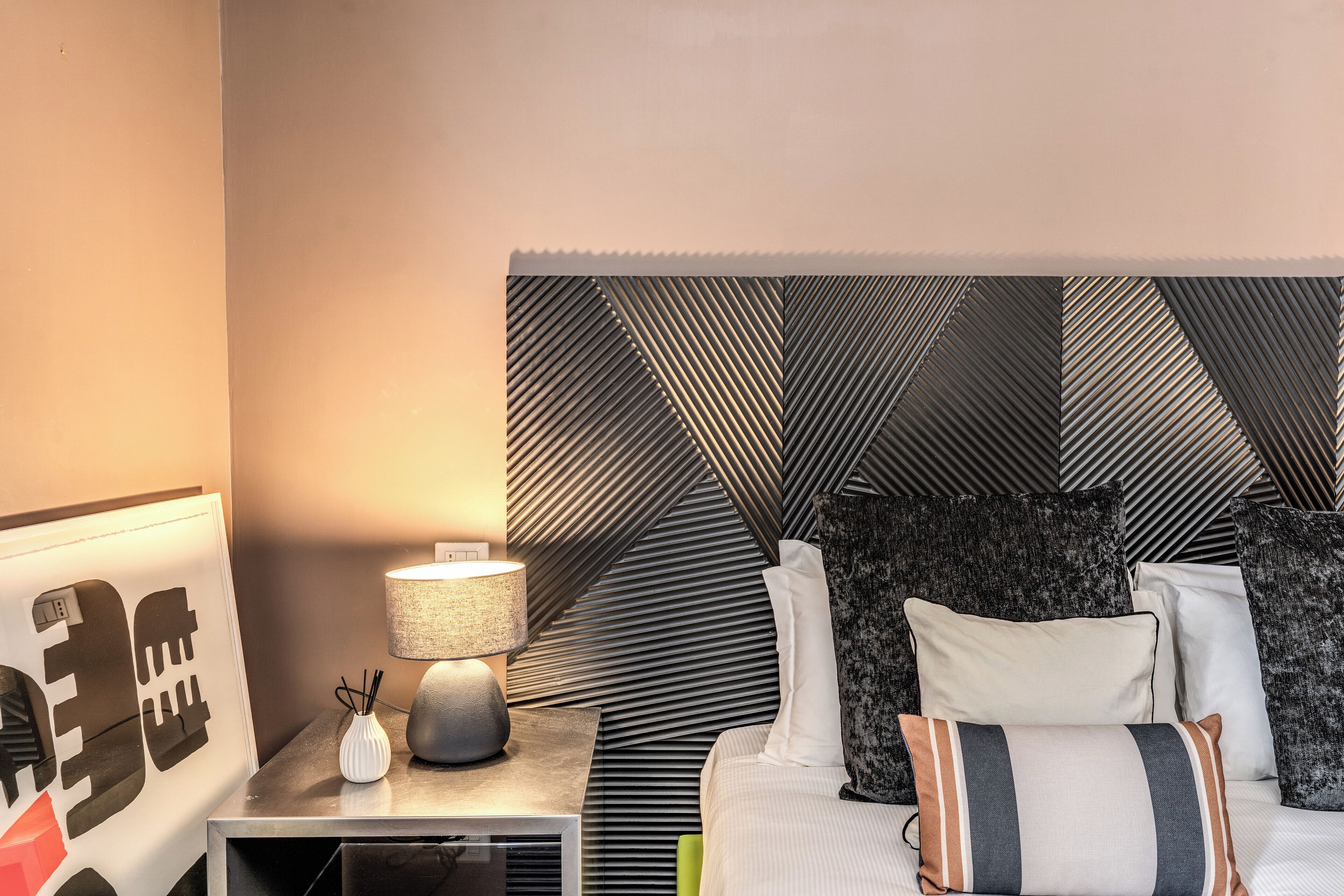 Habitación de hotel impecable con una moderna cabecera, una mesita de noche con lámpara y un cuadro enmarcado.