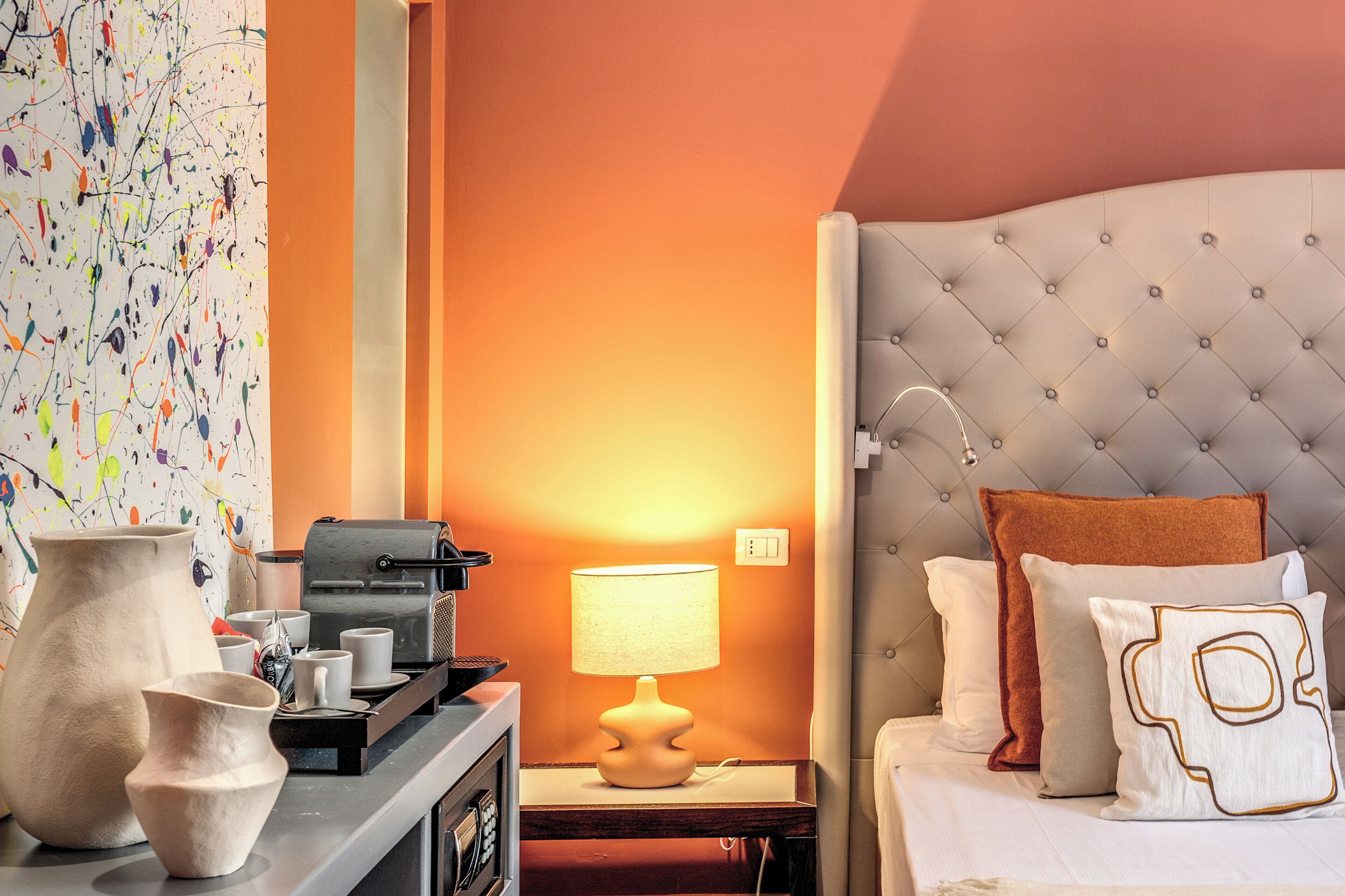 A hotel room with a tufted headboard, a bedside lamp, and a variety of decorative items including vases and a coffee machine.