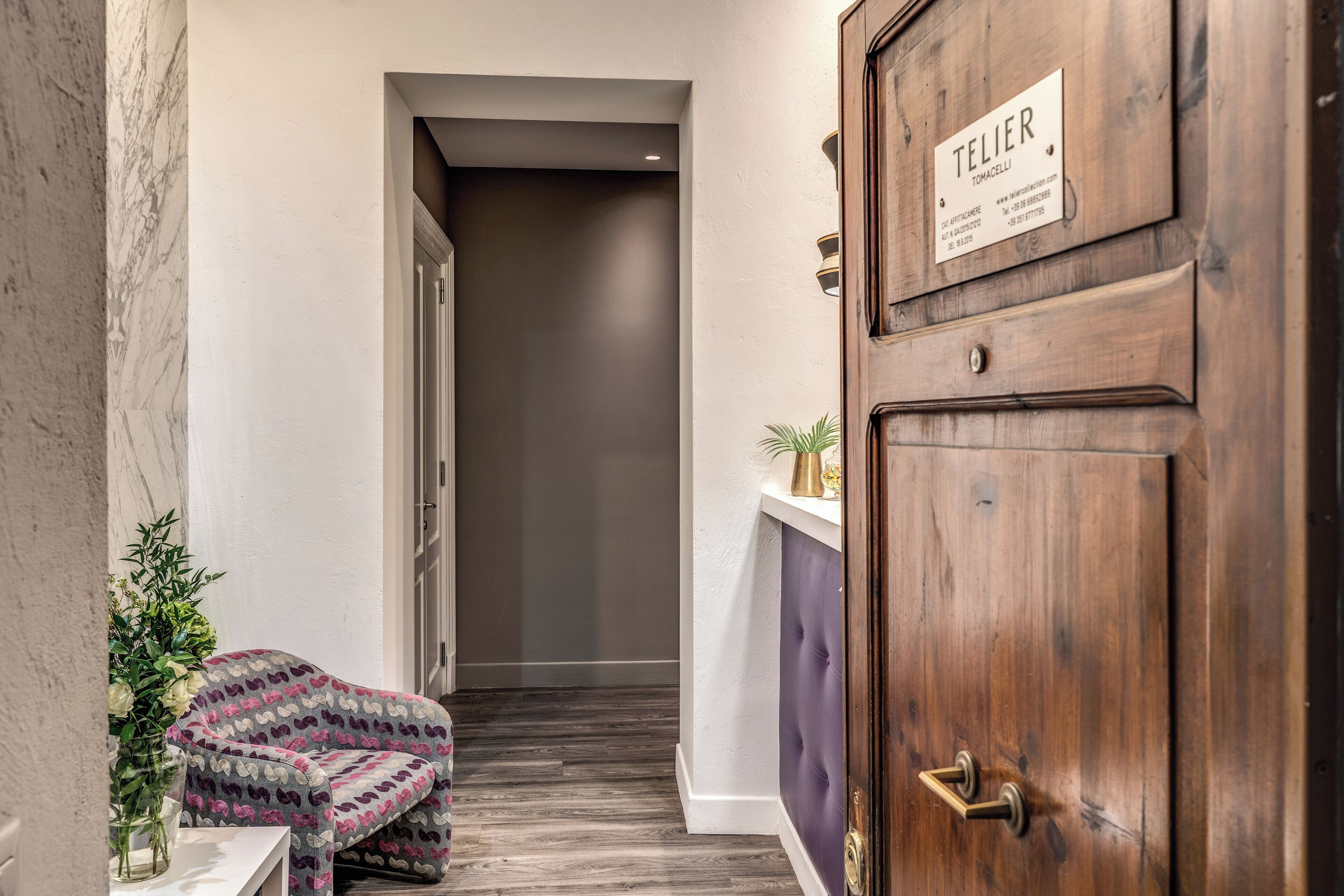 Um corredor com uma porta de madeira com a inscrição 'TELIER', uma poltrona estampada e um vaso com flores.