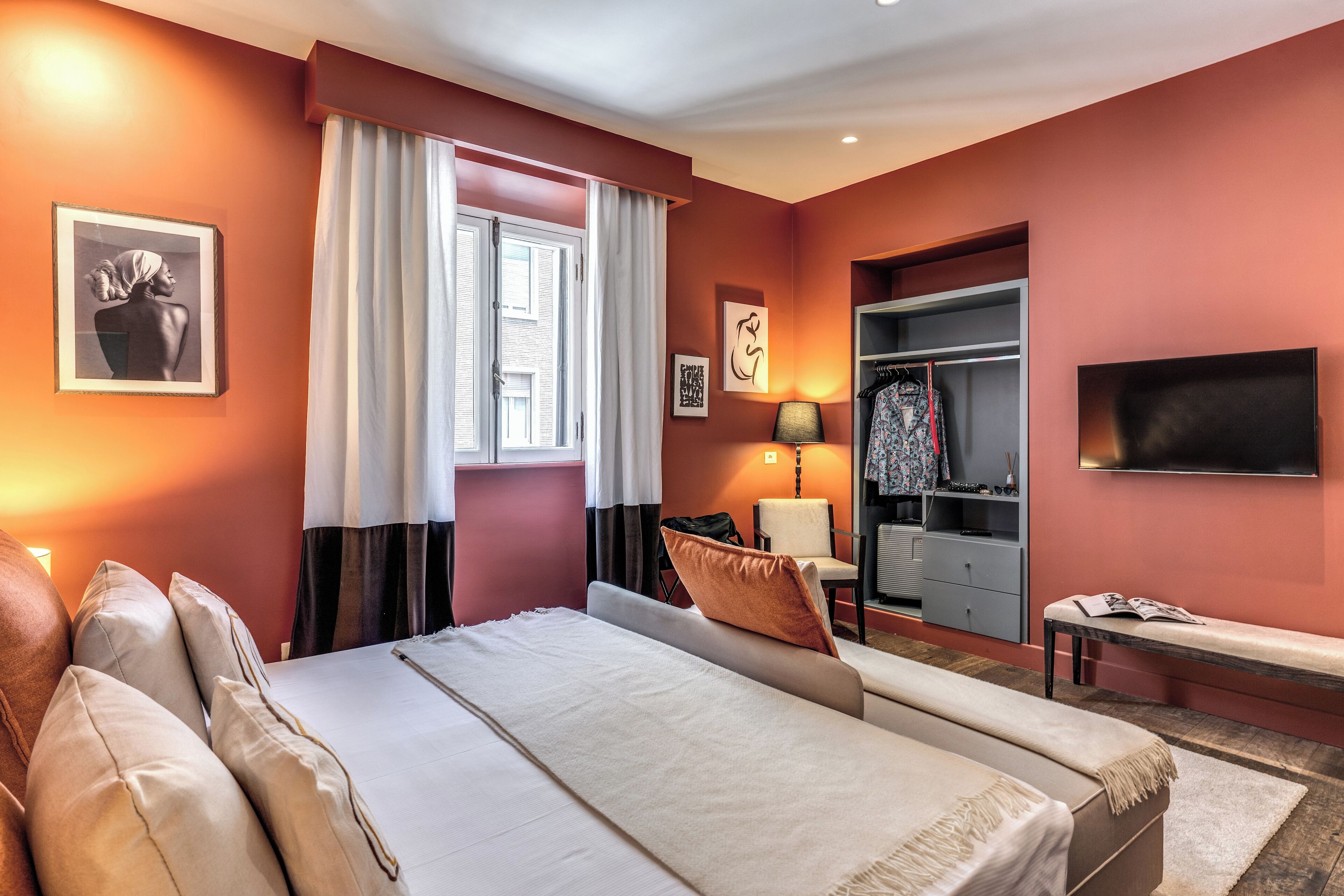 Une chambre d’hôtel avec un lit, une banquette, une télévision, une armoire et une fenêtre avec des rideaux.