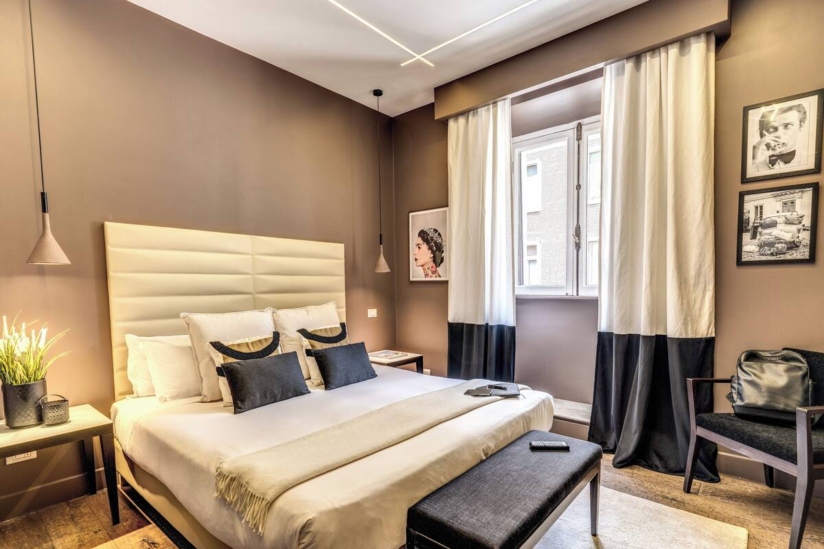 Una habitación de hotel moderna con una cama grande, mesitas de noche, un banco, una silla y una ventana con cortinas.