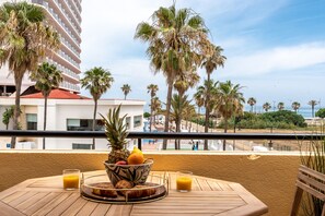 Een balkon met een ontbijttafel, met uitzicht op een strand met palmbomen en een hotel op de achtergrond.