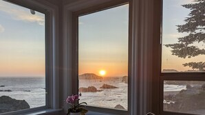 Habitación con un amplio ventanal que ofrece vistas a un atardecer sobre el océano, una planta en maceta en el alféizar y terreno rocoso a la vista.