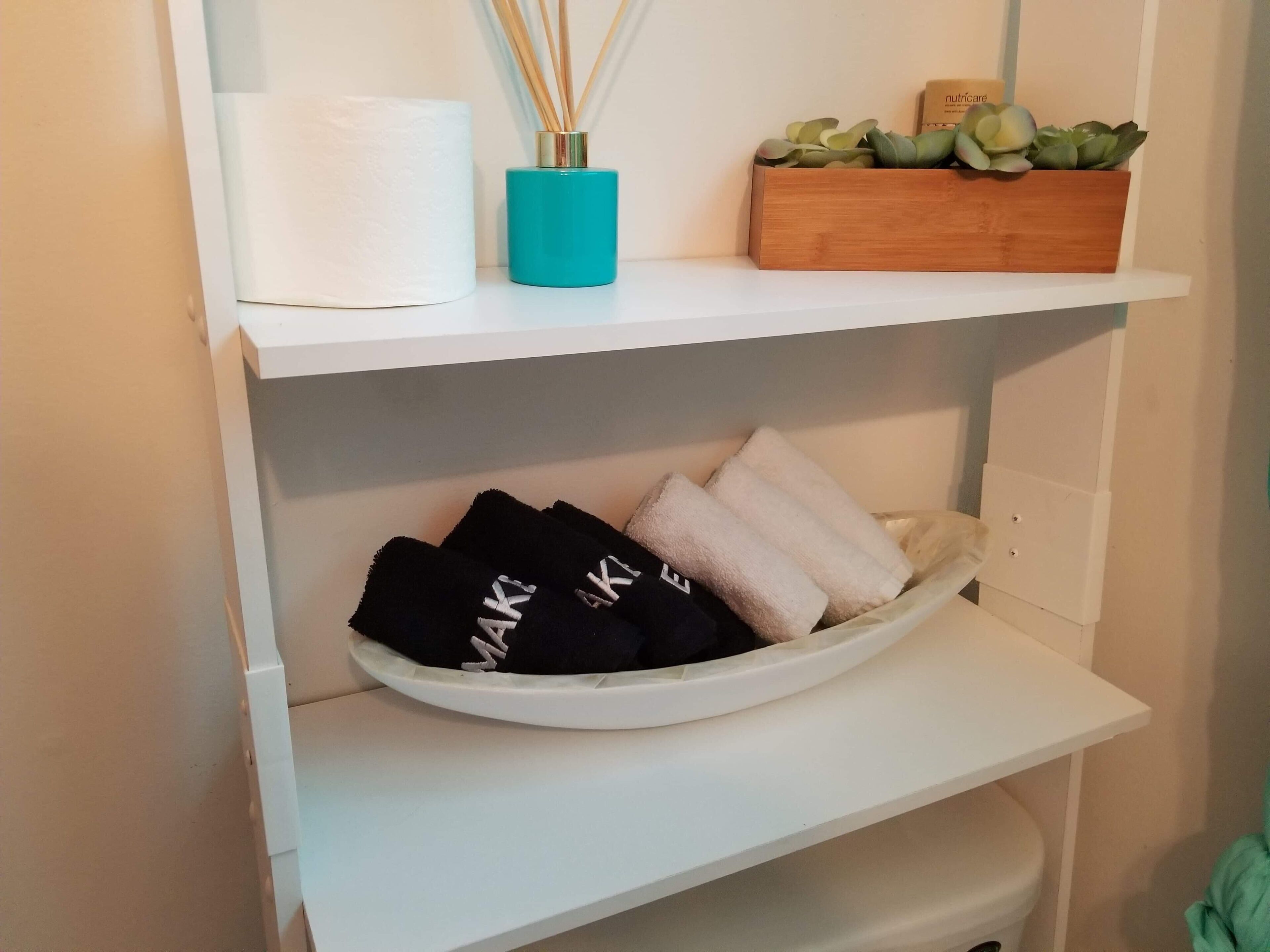 Une étagère de salle de bain avec un rouleau de papier toilette blanc, un vase bleu contenant des diffuseurs à tiges, une boîte en bois avec des plantes succulentes et un bol rempli de serviettes pliées.