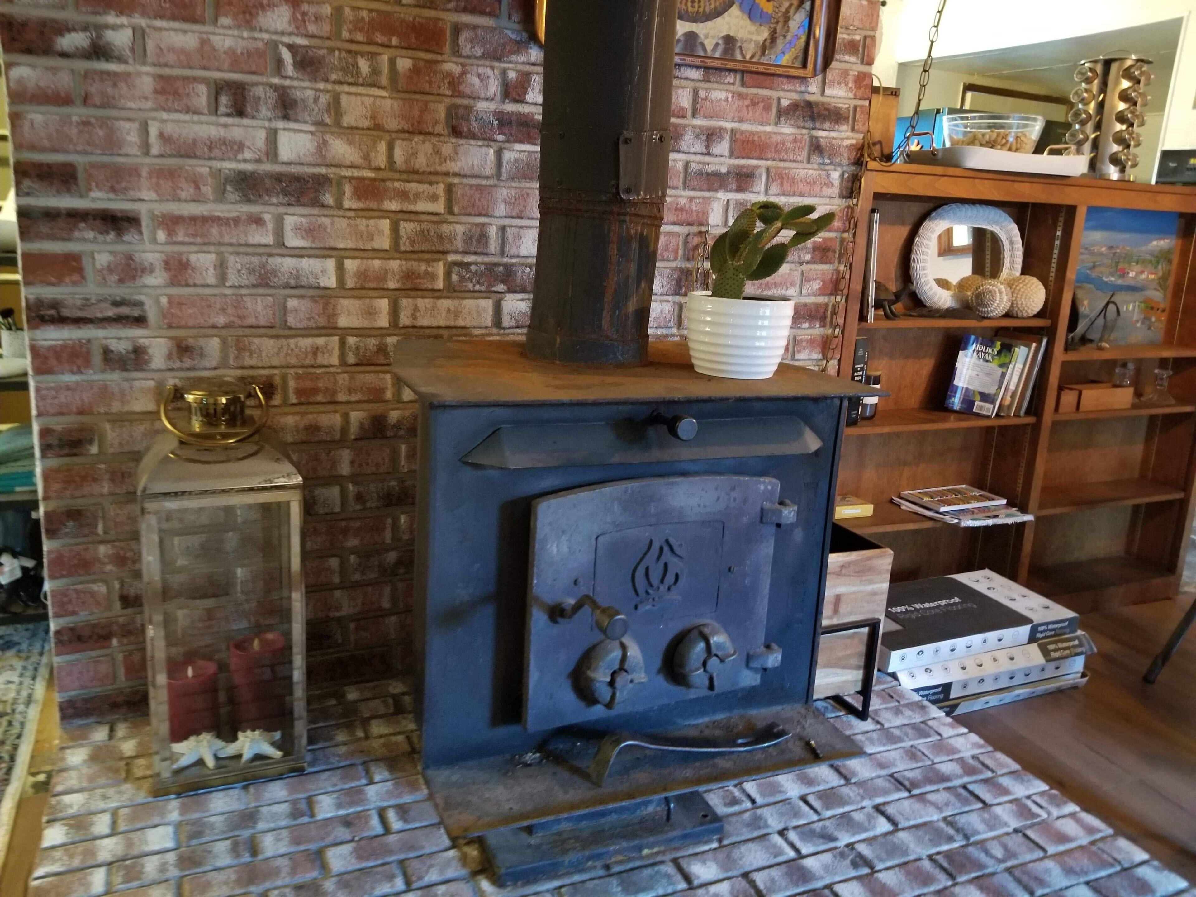 A brick fireplace with a blue stove, a glass vase with a cactus, and a shelf with books and decorative items.