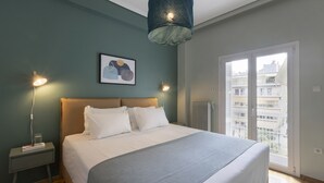 A bedroom with a bed, bedside tables, a lamp, a picture on the wall, and a view through a window. - Volanaki4 - A happy 2 Bdr apartment that has a (Athens)