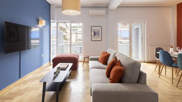 A modern living room with a grey sofa, a red ottoman, and a black coffee table. There is a television mounted on the wall and a dining area with blue chairs. - Volanaki4 - A happy 2 Bdr apartment that has a (Athens)