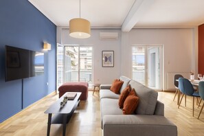Una sala de estar moderna con un sofá gris, un reposapiés rojo y una mesa de centro negra. Hay un televisor montado en la pared y un comedor con sillas azules.
