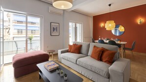 A modern living room with a grey sofa, a red ottoman, and a coffee table with books and plants. There is a dining area with a table set for two, and a balcony with a view of buildings. - Volanaki4 - A happy 2 Bdr apartment that has a (Athens)
