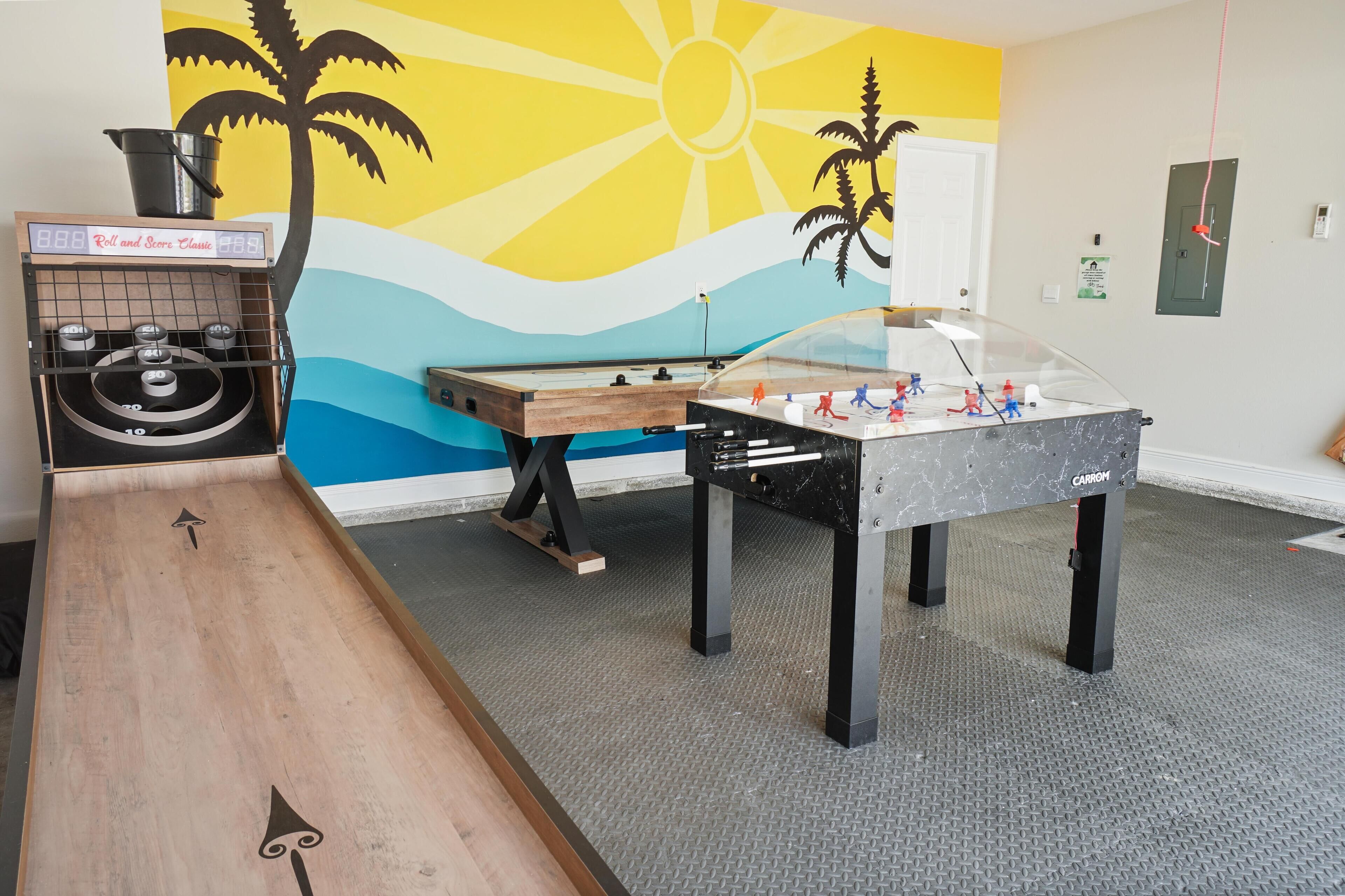 A room with a foosball table, a pool table, and a jukebox against a wall with a tropical mural.