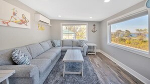 Una sala de estar moderna con un sofá seccional gris, una mesa de centro de madera y un ventanal con vistas a árboles y un edificio.