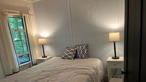 A bedroom with a bed, two bedside tables with lamps, a window with curtains, and a door.
