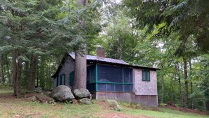 A rustic cabin surrounded by trees, with a large window and a wooden deck.