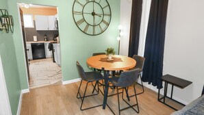Uma área de jantar com uma mesa de madeira redonda, quatro cadeiras, um relógio de parede grande e uma pequena planta.