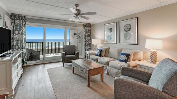 A spacious living room with a large window overlooking the ocean, a ceiling fan, and a flat-screen TV.