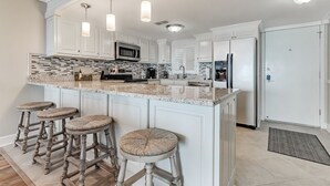 A kitchen with white cabinets, a granite countertop, a microwave, a refrigerator, and a bar stool with a woven seat.