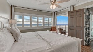 A bedroom with a large bed, a ceiling fan, a view of the ocean through the window, and a door leading to a balcony.