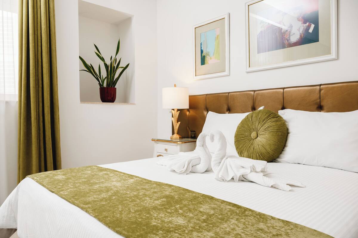 A neatly made bed with a green throw, a potted plant, a nightstand with a lamp, and two framed pictures on the wall.