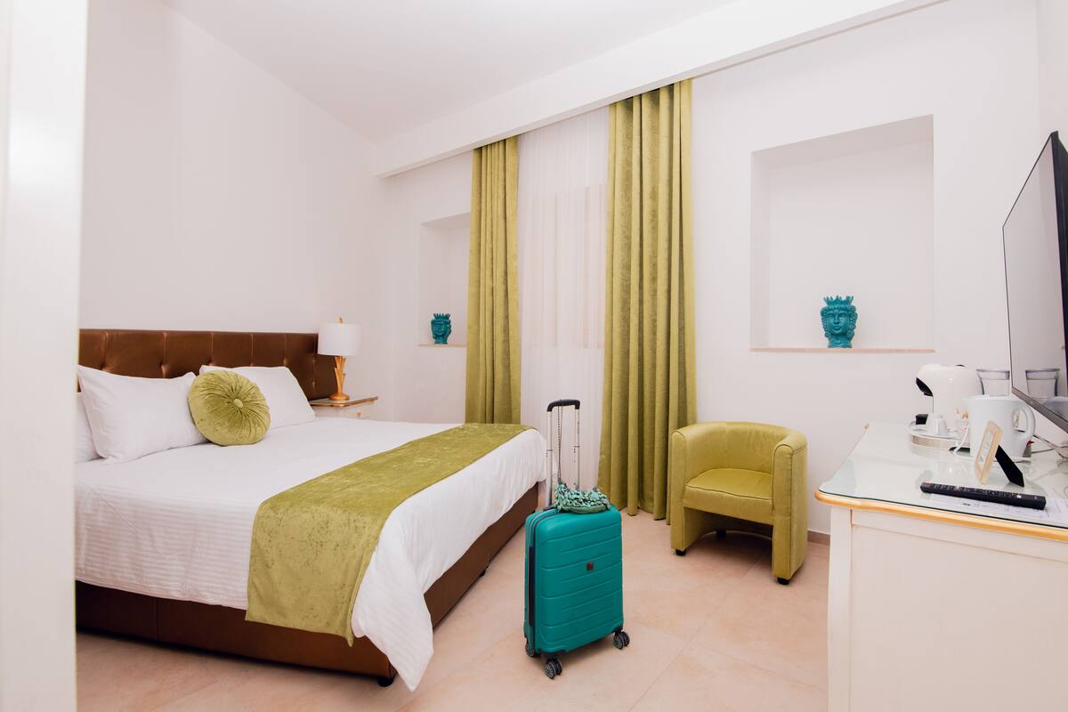 A hotel room with a bed, a green suitcase, a desk with a TV, and a green chair.