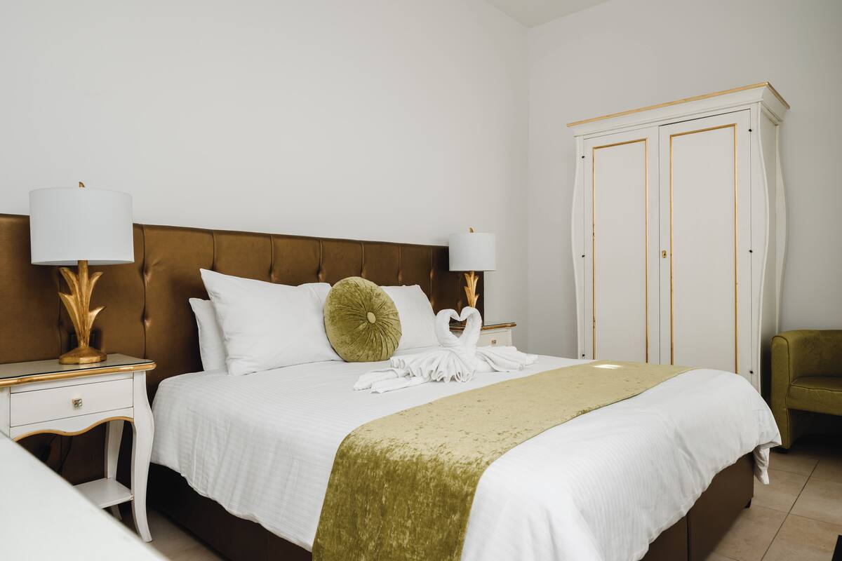 A neatly made bed with white linens, a green throw blanket, and a swan-shaped towel arrangement. There are two bedside tables with lamps, a brown upholstered headboard, and a white wardrobe with gold trim.
